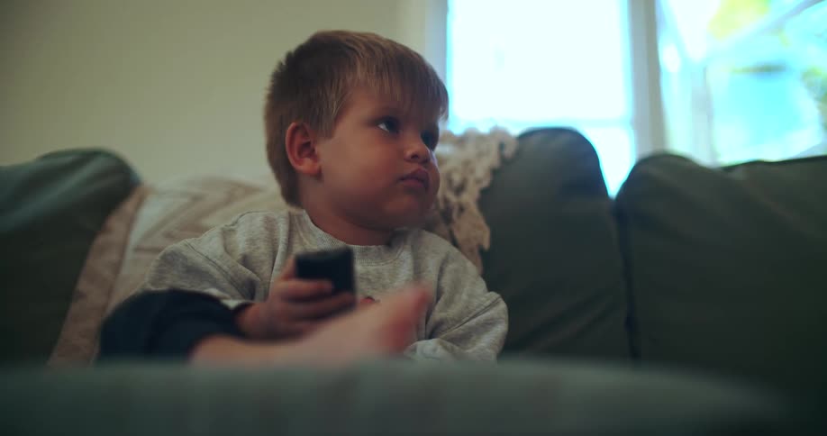 A young boy sits on a green couch, holding a remote control and intently watching television