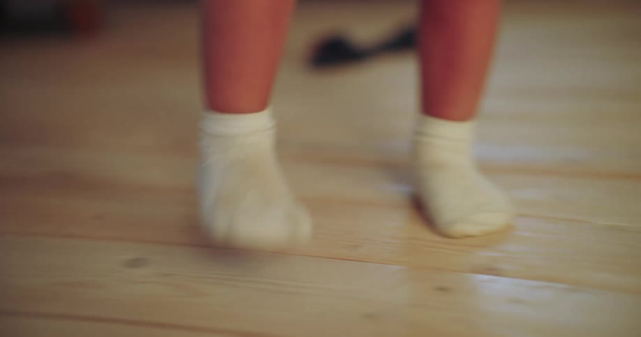 A baby's tiny feet in white socks bounce and move on a light wooden floor as they learn to walk
