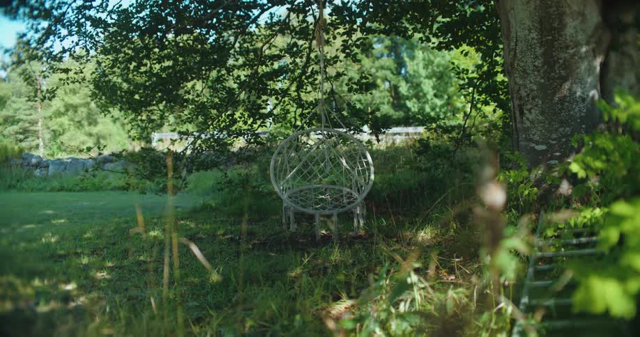 A white macrame hanging chair gently sways in the breeze under the shade of a large tree in a serene garden