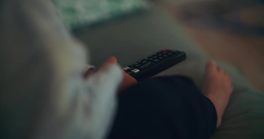 Close-up over-the-shoulder shot of a person relaxing barefoot on a couch while holding a TV remote
