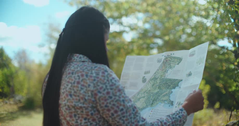 An over-the-shoulder shot of a woman with long hair reading a map in a sunlit park