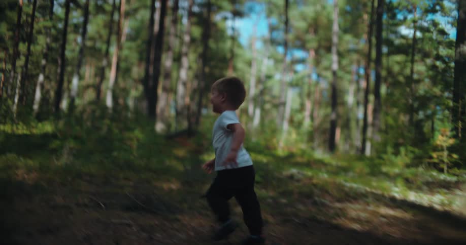 A happy little boy runs with a big smile through a sunny pine forest in slow motion