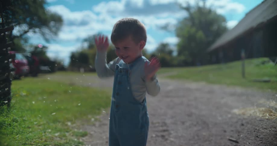 A cute young boy in overalls laughs with pure joy as he throws hay into the air in slow motion on a farm