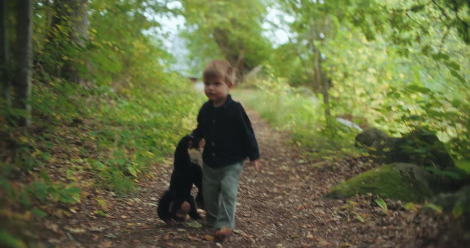 An adorable toddler slowly walks on a forest path, dragging his beloved stuffed monkey by the arm