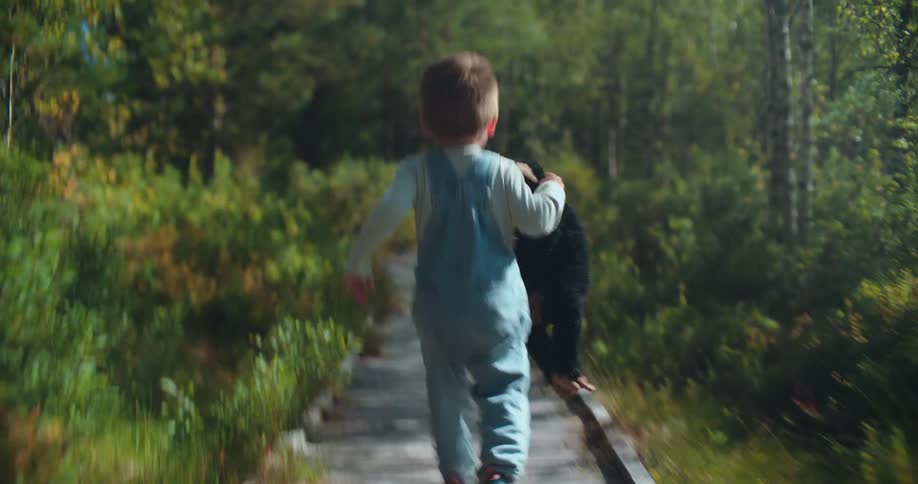 A little boy in overalls runs with his favorite stuffed toy down a wooden boardwalk through a sun-drenched forest