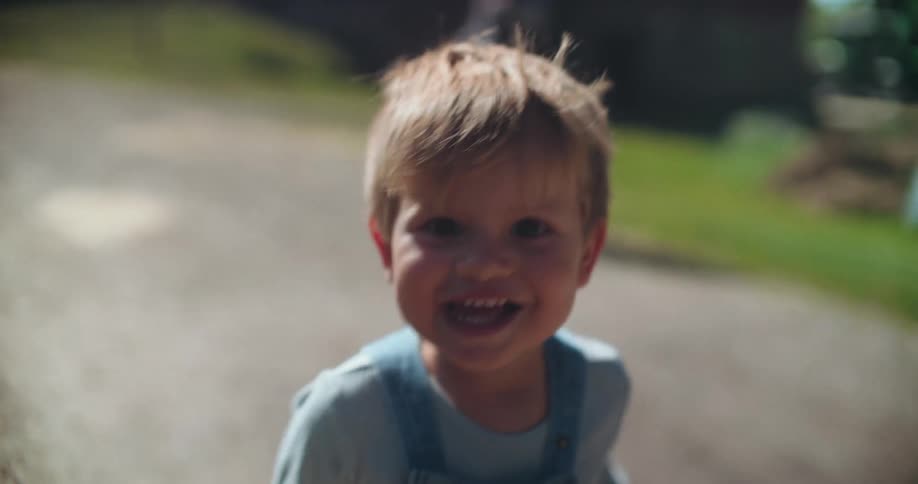 A joyful toddler in overalls runs and laughs towards the camera in slow motion on a sunny day at the farm