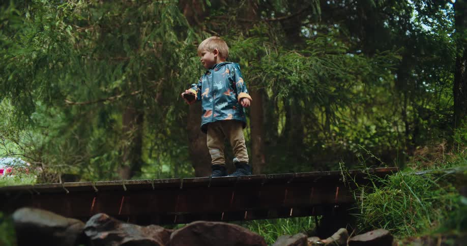 A cute toddler in a raincoat stands on a wooden bridge in the forest and happily drops a rock into the stream below
