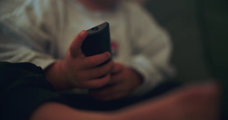 A shallow focus close-up of a young child's hands holding a television remote control in a dimly lit room