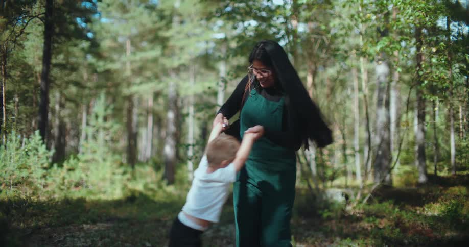 A smiling mother playfully spins her happy young son around in a sun-drenched forest