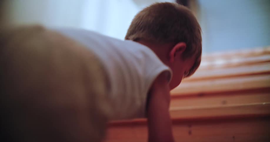 A determined toddler in a white tank top carefully crawls up a wooden staircase in slow motion