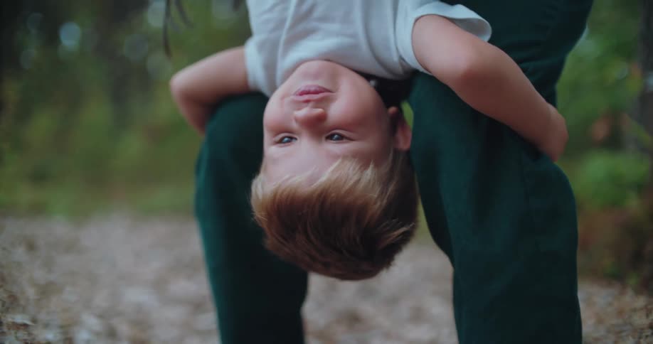 A young boy laughs hysterically while being held upside down by his loving mother in a beautiful forest
