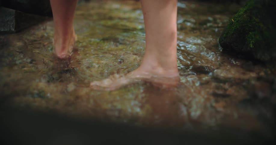 A slow-motion close up of a person's bare feet walking and splashing through a clear, shallow stream in the forest