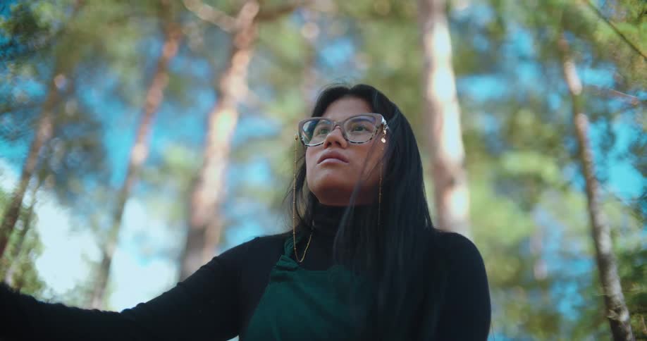 A low-angle shot of a woman with glasses looking up contemplatively in a sunlit pine forest