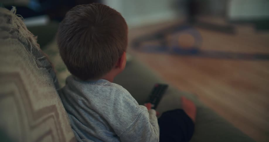 Over the shoulder shot of a toddler sitting on a couch and holding a remote control while watching television