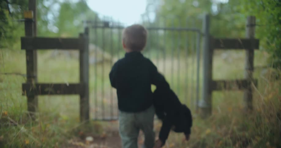 A small boy carrying a large stuffed animal companion walks down a leafy path and tries to open a locked gate in slow motion