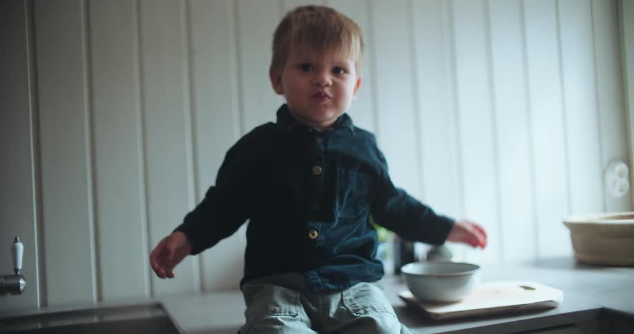 A cute toddler with a full mouth sits on the kitchen counter eating a snack from a bowl and makes a funny face