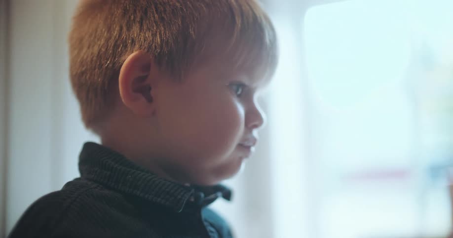 Close-up slow-motion shot of a cute little boy with a curious expression looking out of a bright window