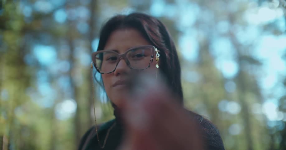 A woman with glasses in a tranquil forest holds a small pinecone, smelling its earthy scent in a slow-motion close-up