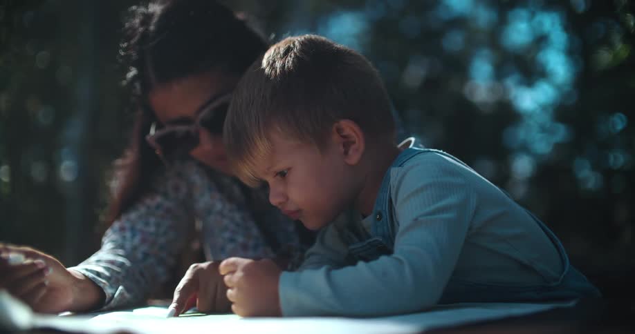 A curious little boy intently studies a trail map with his mother in a sunlit forest, making a funny pouty face