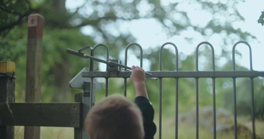 A determined young boy reaches up to try and open a metal gate on a wooded trail in slow motion