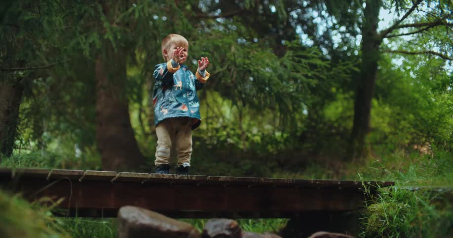 A curious toddler in a raincoat gleefully drops a rock from a wooden bridge into a forest stream, creating a splash