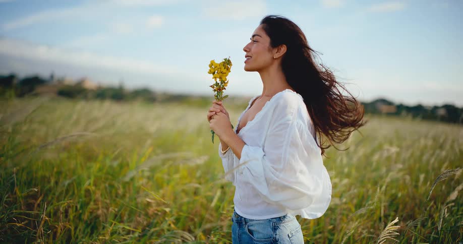 A peaceful woman with long dark hair holds yellow flowers and enjoys the wind blowing through a field of tall grass