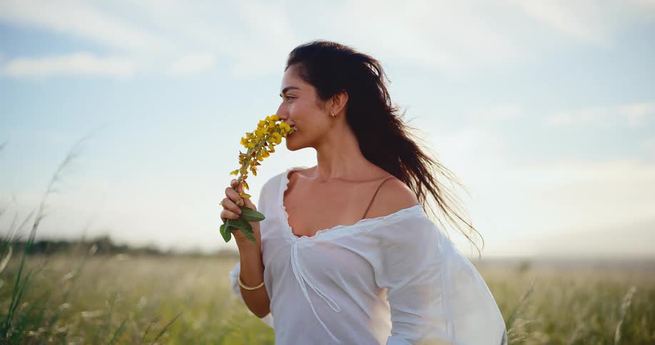 A beautiful young woman smells a bouquet of yellow wildflowers in a sun-drenched grassy field in slow motion