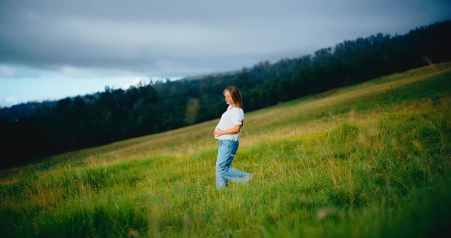 A happy pregnant woman walks through a lush green field on a hillside, lovingly cradling her belly in slow motion