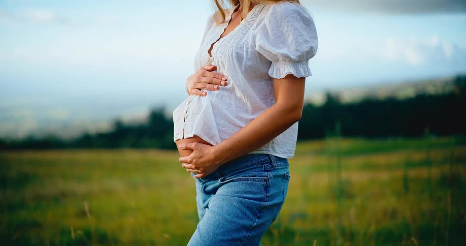 A pregnant woman in a white blouse and jeans gently holds her baby bump while standing in a beautiful green field