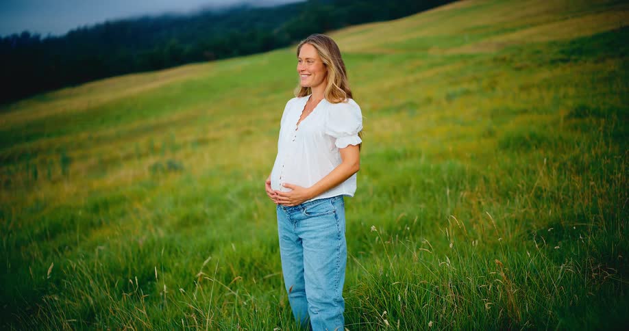 A happy pregnant woman gently holds her belly while standing and smiling in a beautiful green field in slow motion