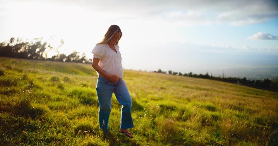 Joyful pregnant woman lovingly holds her belly as she walks through a golden, sun-drenched grassy meadow at sunset