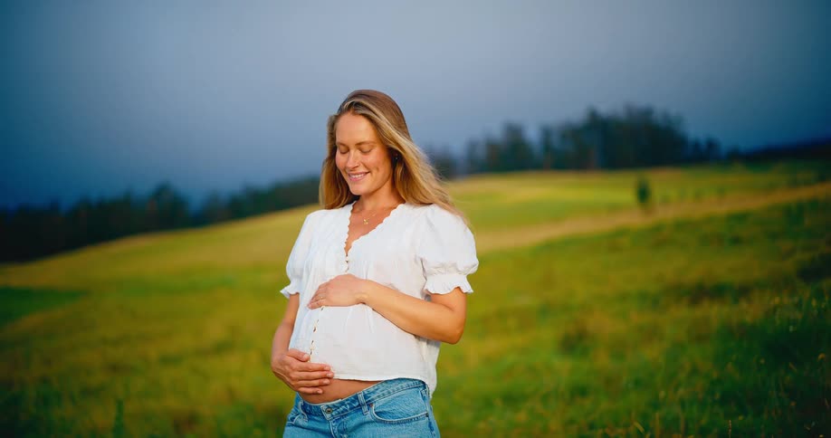 Beautiful happy pregnant woman smiling and holding her pregnant belly in a green field with her hair blowing in the wind