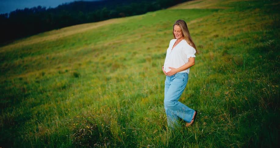 Radiant pregnant woman walks peacefully through a lush green field, gently cradling her baby bump in slow motion