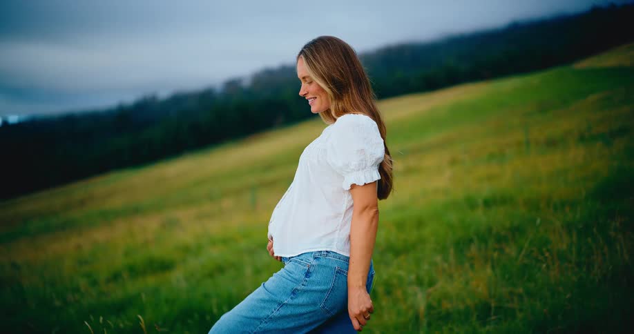 A beautiful pregnant woman smiles peacefully as she holds her baby bump while standing in a lush green field at dusk
