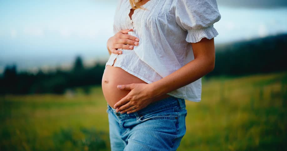 A pregnant woman gently holds her bare belly while standing in a beautiful green field