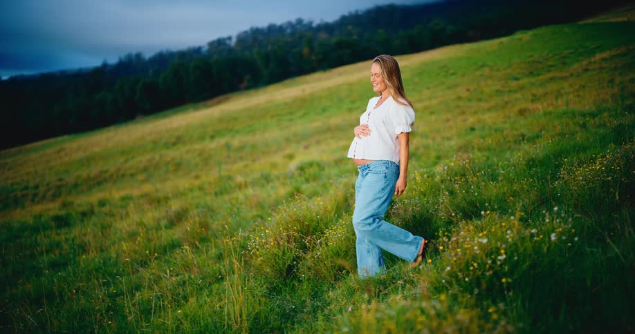 A happy pregnant woman gently holds her belly while walking through a beautiful green field at dusk