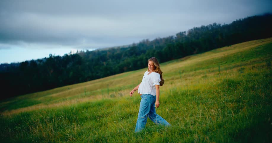 A beautiful pregnant woman smiles and walks through a lush green field in slow motion at dusk