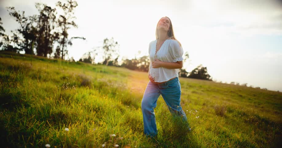 A serene pregnant woman gently holds her belly while walking slowly up a beautiful grassy hill in the bright afternoon sun