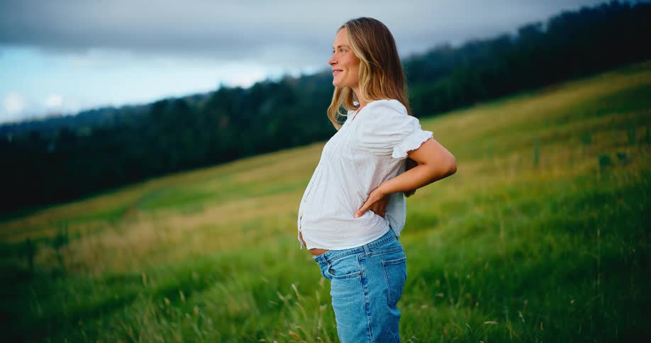 A serene pregnant woman lovingly holds her baby bump while standing in a beautiful green field at dusk