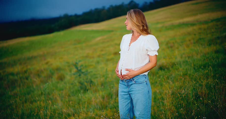 A beautiful pregnant woman smiles peacefully while holding her baby bump in a vast green field at dusk