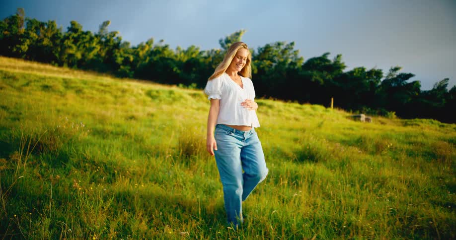 A beautiful smiling pregnant woman walks through a sunlit grassy field cradling her belly in slow motion