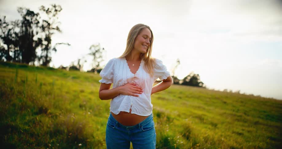 A beautiful pregnant woman gently holds her belly while standing in a sun-drenched green field, smiling with joy