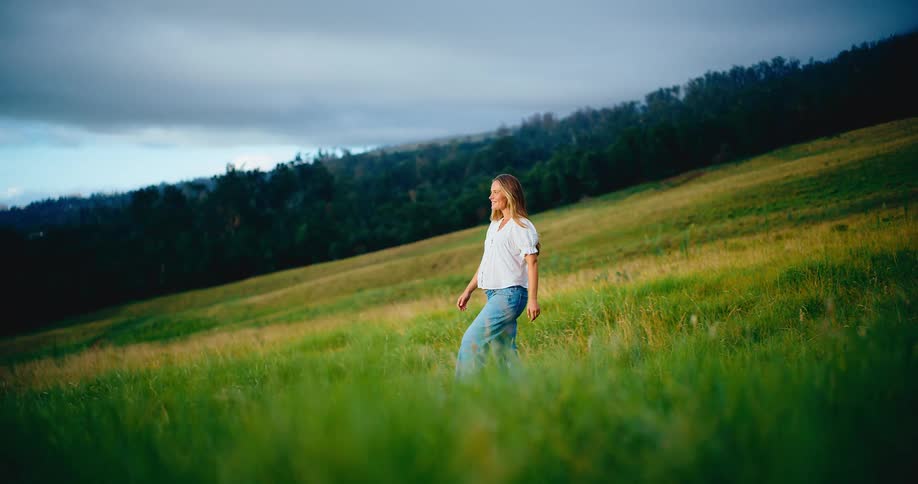 A beautiful pregnant woman smiles and walks through a picturesque green field in slow motion at sunset