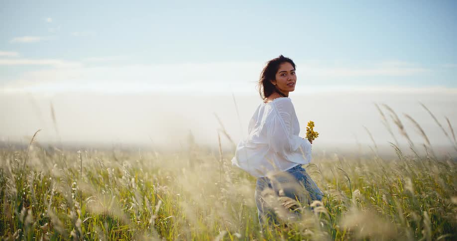 A serene young woman with a bouquet of wildflowers looks back while standing in a breezy, sun-drenched field of tall grass