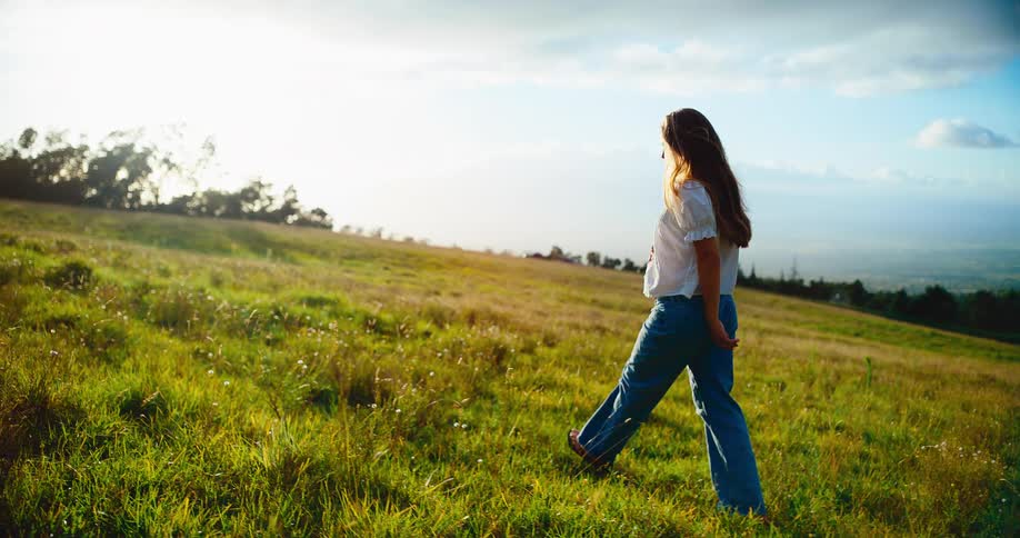 A radiant pregnant woman strolls peacefully through a vast green field during a golden sunset in slow motion