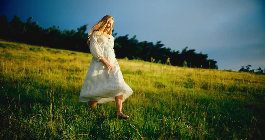In slow motion, a beautiful pregnant woman in a flowing white dress walks joyfully through a golden sunlit meadow