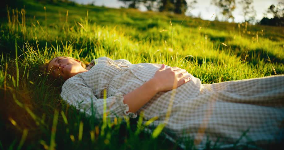 A peaceful pregnant woman rests in a lush green meadow, basking in the warm golden hour sunlight with her hand on her belly