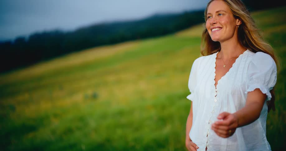 A happy pregnant woman wearing a white top smiles peacefully while walking through a beautiful green field at sunset
