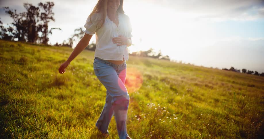 A radiant pregnant woman enjoys a peaceful walk through a golden, sunlit meadow at sunset