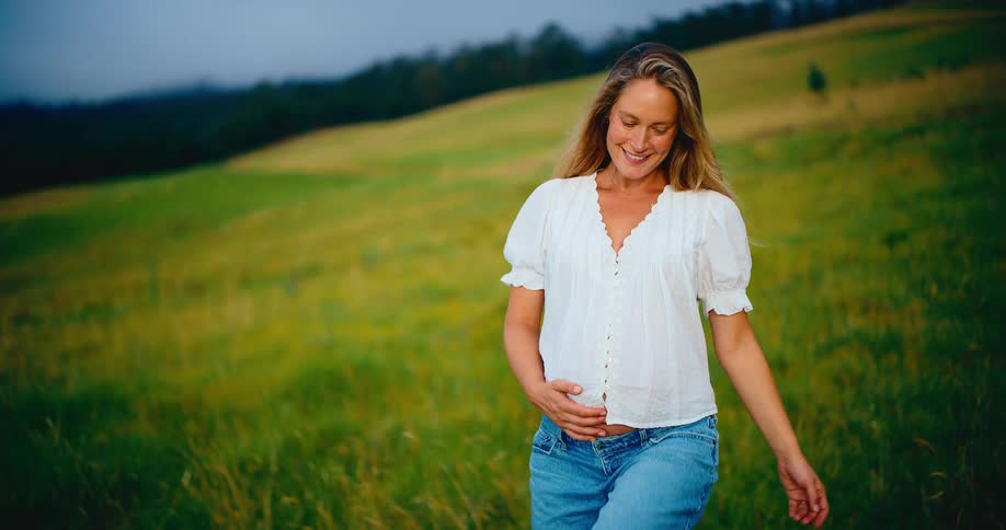 A serene pregnant woman enjoys a peaceful walk in a beautiful green field, cradling her baby bump